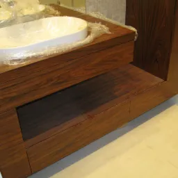 Nowoczesna umywalka nablatowa, jeszcze zabezpieczona folią, na blacie z ciemnego drewna egzotycznego z widoczną strukturą słojów, w łazience z jasnymi płytkami i kabiną prysznicową.