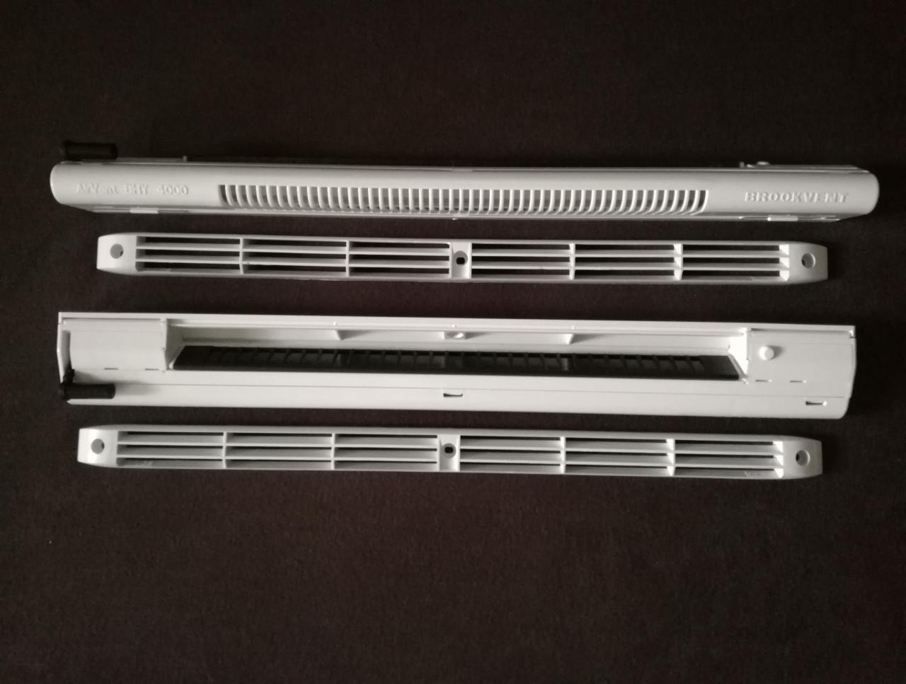 Białe nawiewniki okienne Brookvent i A-Vent PHY 4000, ułożone równolegle na ciemnym tle, widoczne kratki wentylacyjne i detale konstrukcyjne.