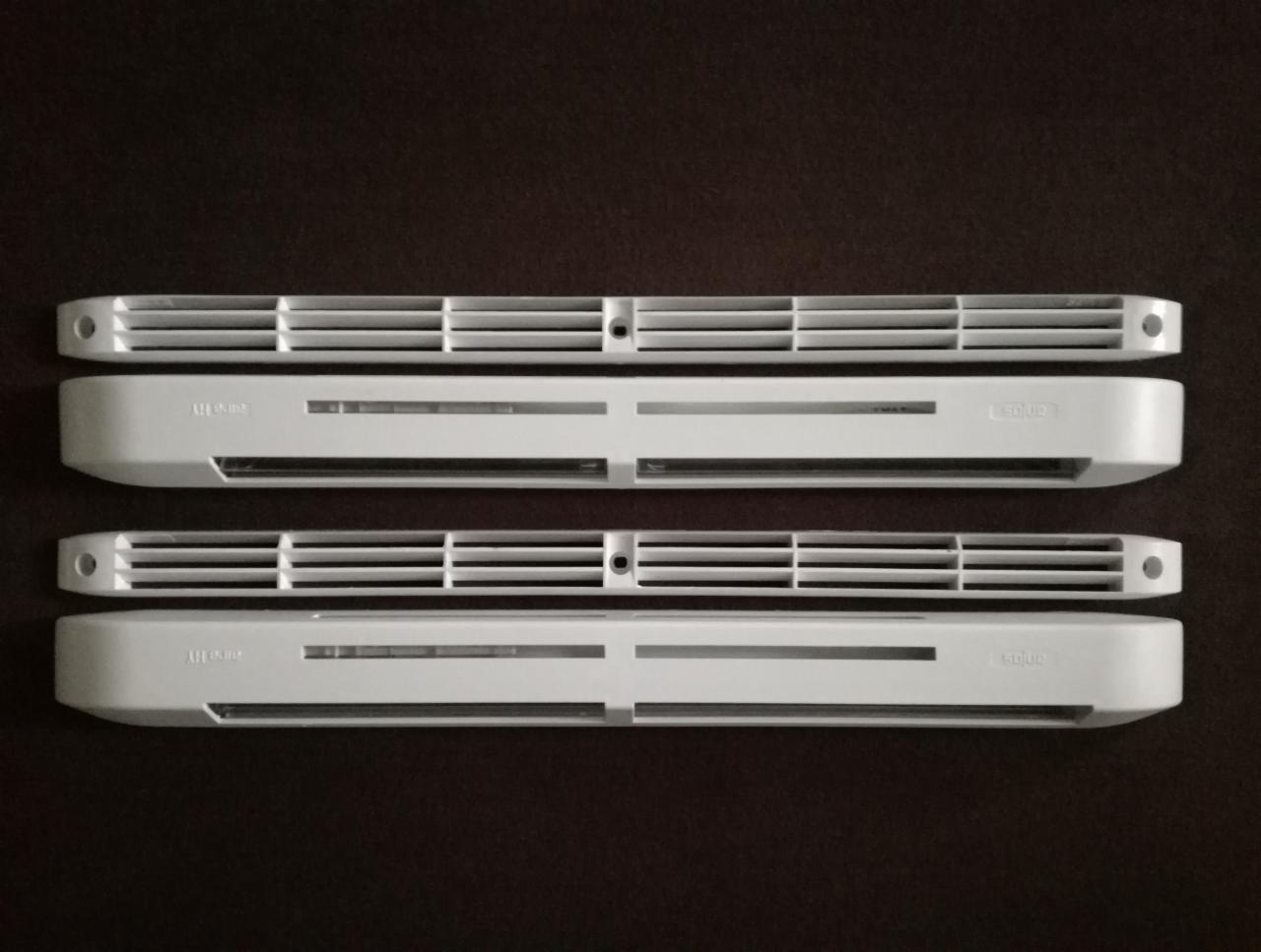 Dwa białe, prostokątne nawiewniki wentylacyjne z kratkami, ułożone jeden nad drugim na brązowym tle, widoczne otwory montażowe i oznaczenia modelu.