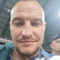 Mężczyzna z krótką fryzurą i lekkim zarostem, ubrany w polo z niebiesko-białymi pasami, pozuje na tle hali sportowej z widocznymi trybunami i oświetleniem.