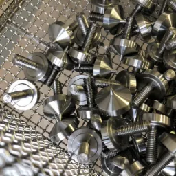 Partie metalowych, precyzyjnie obrobionych elementów z gwintem i stożkową główką, leżących w metalowym koszu o drobnych oczkach.