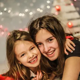 Dwie roześmiane dziewczynki, w tym jedna z pomalowanymi na niebiesko paznokciami, przytulają się na tle rozmytych światełek w Rokitnicy. Młodsza dziewczynka ubrana jest w czerwoną bluzę.