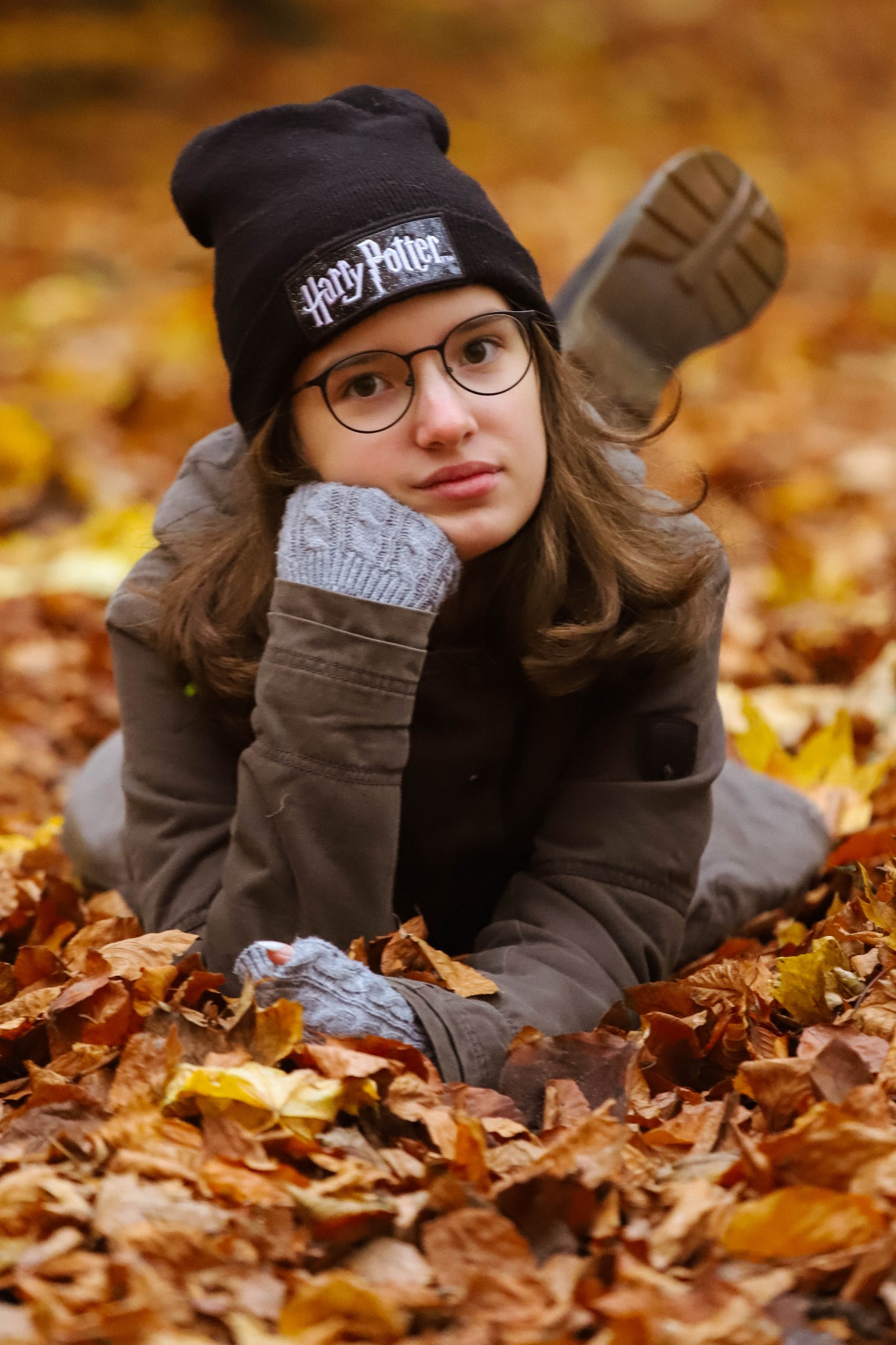 Młoda osoba w czapce z napisem Harry Potter, okularach i szarych rękawiczkach leży na opadłych, brązowo-żółtych liściach jesienią.