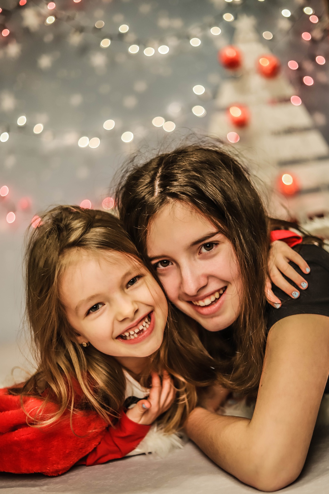 Dwie roześmiane dziewczynki, w tym jedna z pomalowanymi na niebiesko paznokciami, przytulają się na tle rozmytych światełek w Rokitnicy. Młodsza dziewczynka ubrana jest w czerwoną bluzę.