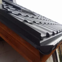 Fragment dachu pokrytego blachą w kolorze grafitowym, z zamontowaną rynną w tym samym kolorze, widoczne krople wody po deszczu.