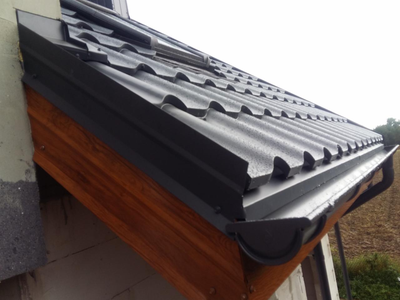 Fragment dachu pokrytego blachą w kolorze grafitowym, z zamontowaną rynną w tym samym kolorze, widoczne krople wody po deszczu.