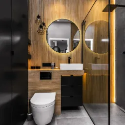 Nowoczesna łazienka z drewnianymi panelami, okrągłym lustrem z podświetleniem LED, czarną armaturą i szklaną kabiną prysznicową.