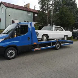 Niebieska laweta Iveco z białym, klasycznym Mercedesem na platformie, zaparkowana na brukowanej ulicy przed domem.
