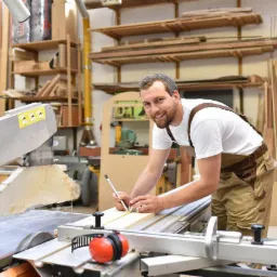 Uśmiechnięty mężczyzna w roboczym stroju mierzy deskę na stole z piłą tarczową w warsztacie, na półkach widoczne zapasy drewna.