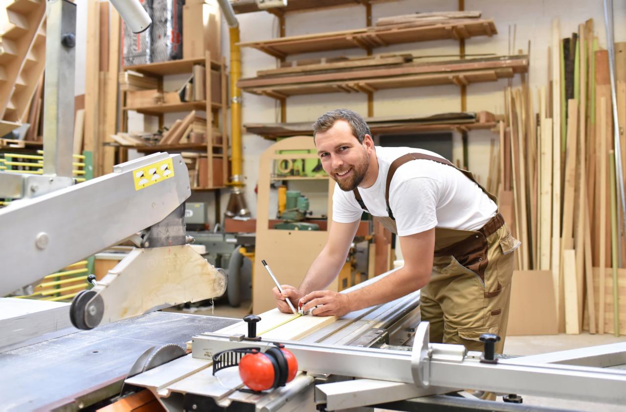 Uśmiechnięty mężczyzna w roboczym stroju mierzy deskę na stole z piłą tarczową w warsztacie, na półkach widoczne zapasy drewna.