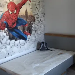 Szare łóżko z drewnianymi elementami w pokoju z fototapetą Spidermana na ścianie, materac owinięty folią ochronną, plecak leży na łóżku.