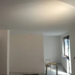 Wnętrze w trakcie remontu: widoczne świeżo pomalowane ściany na biało, okno z zasłoniętą ramą, metalowe krzesło i drabina, przewody zwisające z sufitu, oraz gniazdko elektryczne.