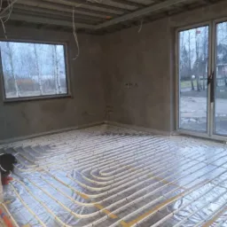 Surowy stan pomieszczenia z widoczną instalacją ogrzewania podłogowego na siatce, przygotowaną do wylewki, szare ściany, sufit z metalową konstrukcją, okno z widokiem na drzewa.