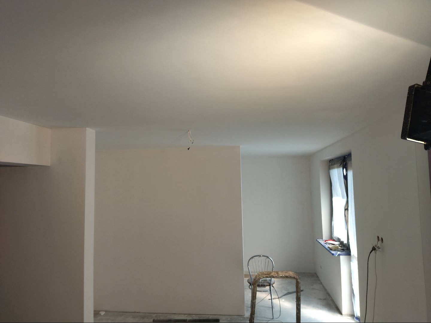 Wnętrze w trakcie remontu: widoczne świeżo pomalowane ściany na biało, okno z zasłoniętą ramą, metalowe krzesło i drabina, przewody zwisające z sufitu, oraz gniazdko elektryczne.
