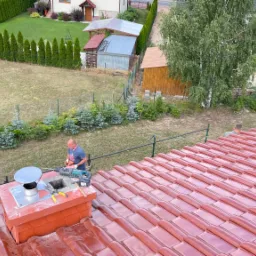 Czerwony dach z dachówki ceramicznej, komin z metalowym wkładem, dekarz z wiertarką na tle zielonego ogrodu z tujami i budynkami gospodarczymi.