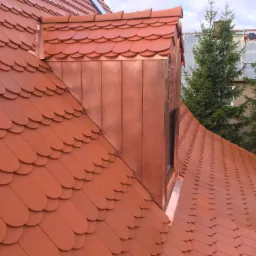 Nowy dach pokryty dachówką karpiówką w kolorze ceglastym, z miedzianą obróbką komina i lukarną, na tle zielonych drzew.