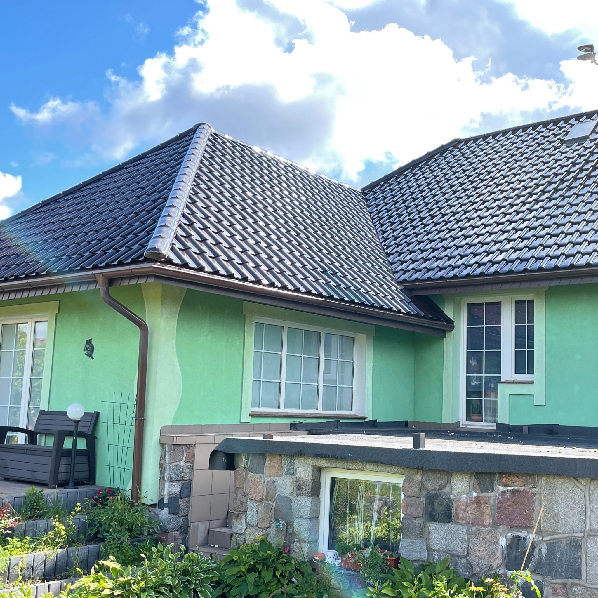 Dom z zieloną elewacją i nowym, ciemnym dachem z dachówki ceramicznej, brązowe rynny, widoczne okna i kamienna podmurówka.