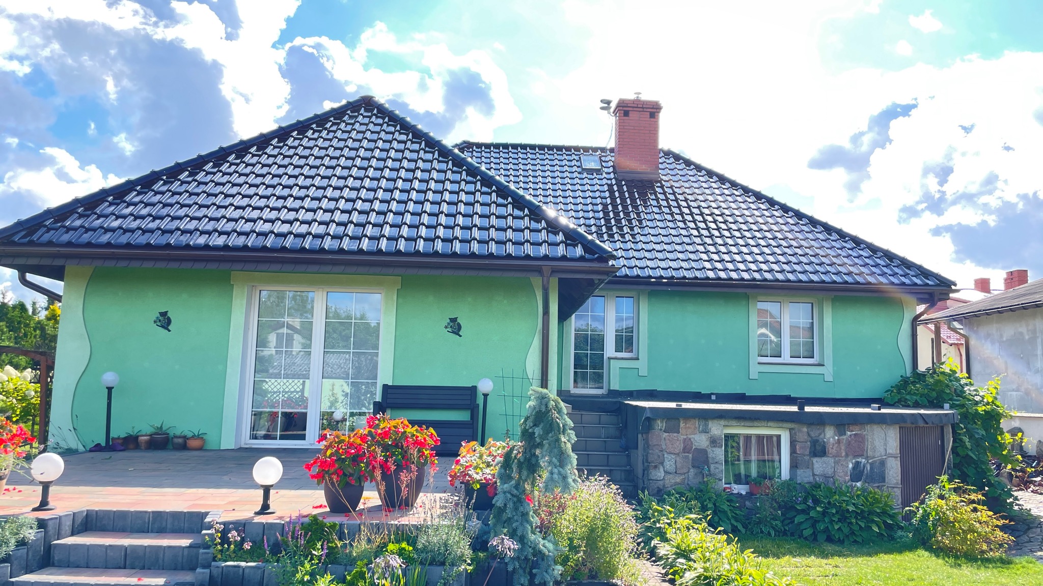 Dom jednorodzinny z ciemną dachówką ceramiczną, zieloną elewacją z dekoracyjnym motywem i zadbanym ogrodem z kwiatami.
