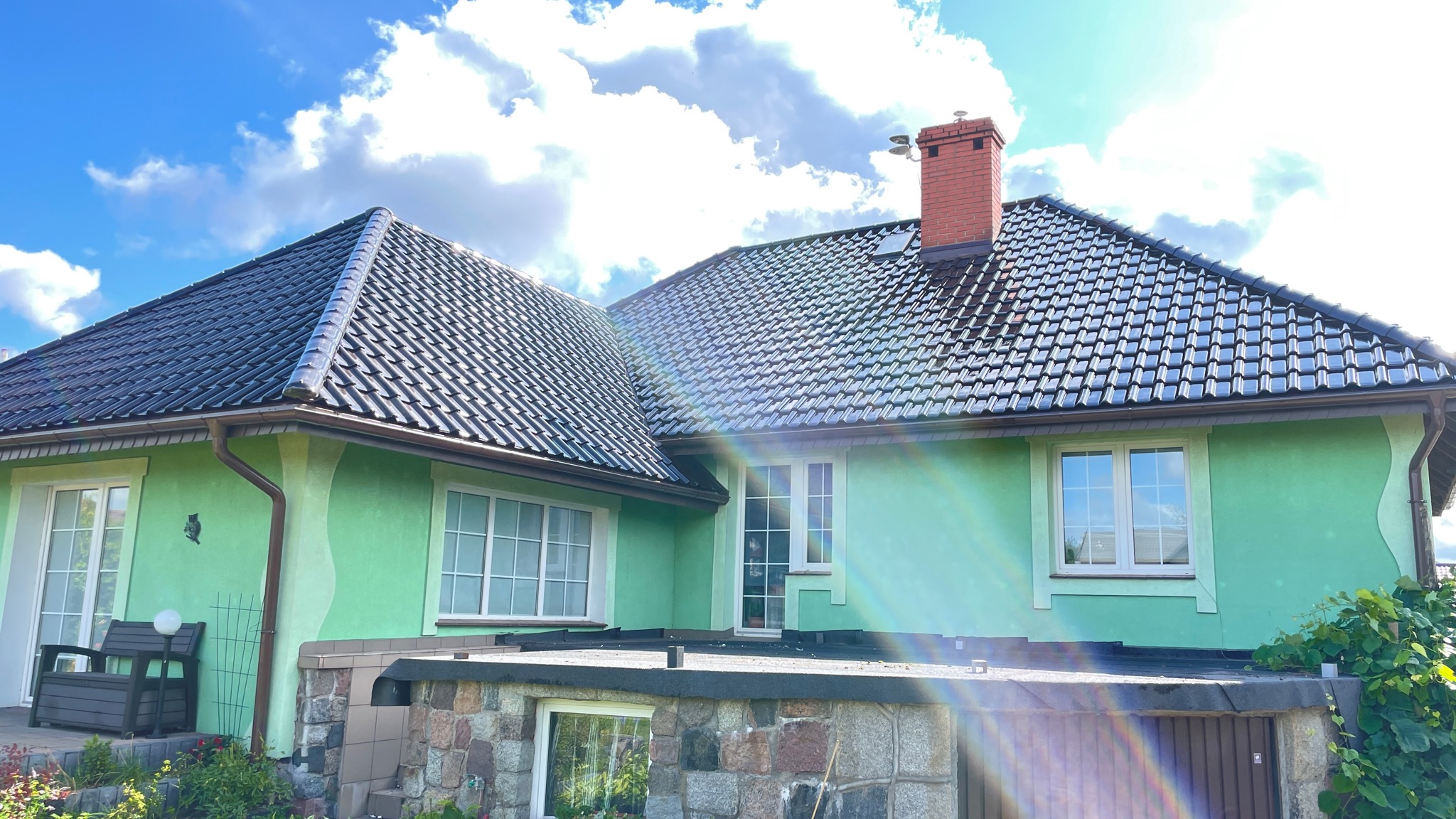 Dom z zieloną elewacją i błyszczącym, ciemnym dachem ceramicznym w słoneczny dzień, widoczny komin z cegły i system rynnowy.