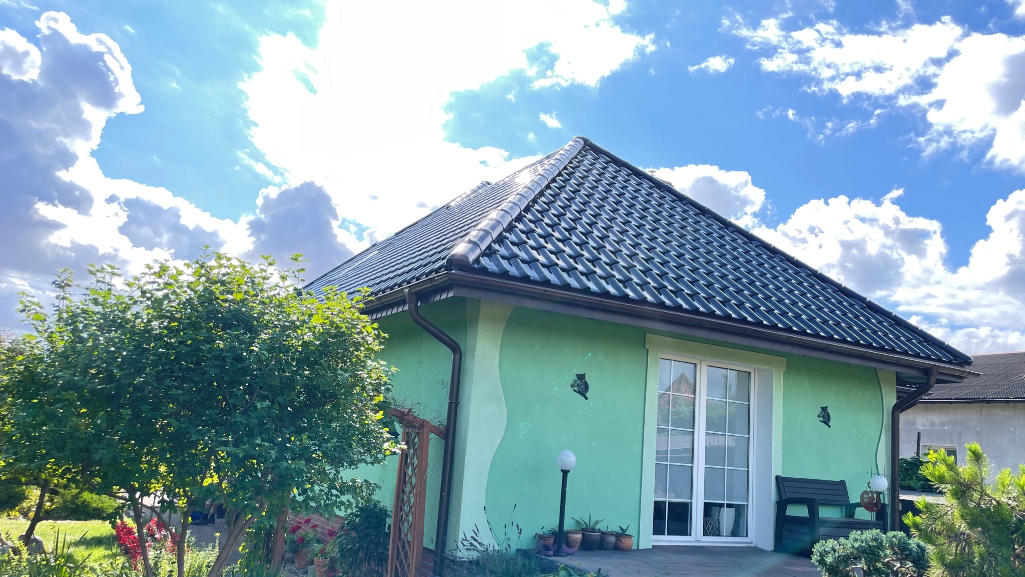 Dom z zieloną elewacją i ciemnym dachem z dachówki ceramicznej, widoczne rynny, fragment ogrodu oraz dekoracja ścienna w kształcie sowy.