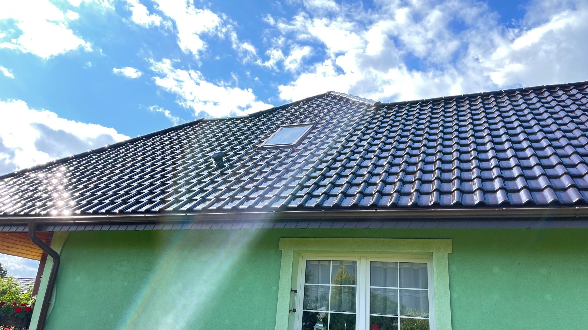 Błyszczący, ciemny dach z dachówki ceramicznej z oknem dachowym, widoczny fragment zielonej elewacji z oknem i fragment systemu rynnowego, na tle błękitnego nieba z chmurami.