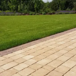 Ułożona z beżowej kostki brukowej ścieżka w ogrodzie, przylegająca do idealnie przystrzyżonego trawnika z rabatą lawendy w tle.