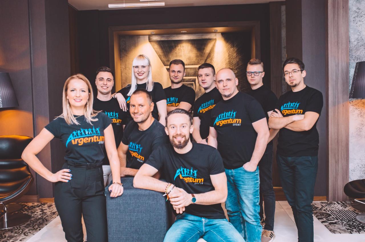 Zespół w czarnych koszulkach z logo firmy eventowej, pozujący w eleganckim wnętrzu z abstrakcyjnym obrazem w tle.