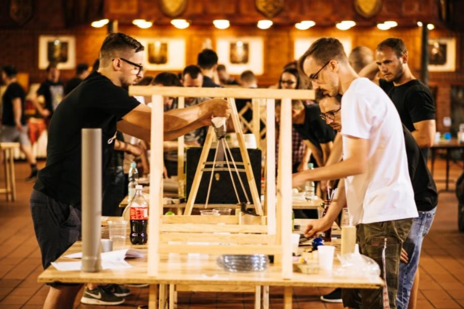 Grupa mężczyzn w okularach, pochylonych nad drewnianymi konstrukcjami na stołach, prawdopodobnie podczas warsztatów budowlanych lub team buildingu.