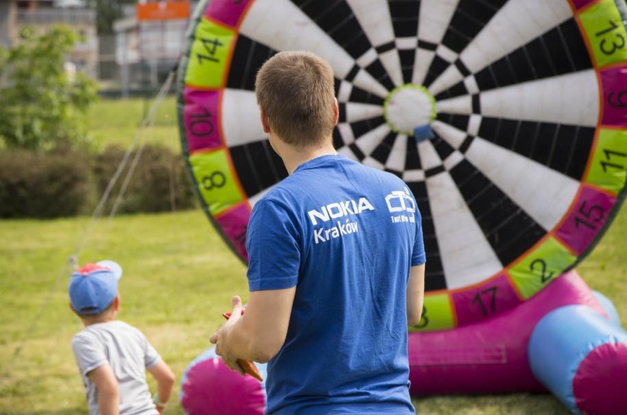 Mężczyzna w niebieskiej koszulce z logo Nokia Kraków rzuca lotkami do dmuchanej tarczy z kolorowymi polami punktowymi podczas pikniku firmowego na trawniku.