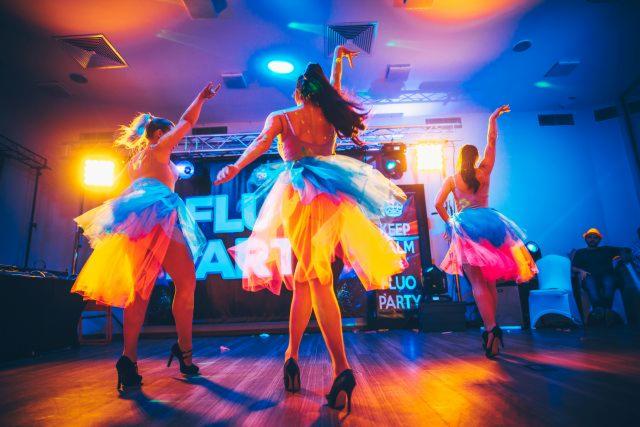 Trzy tancerki w kolorowych tutu na scenie pod światłami, z napisem 'Fluo Party' w tle, widziane z tyłu, na wysokich obcasach.