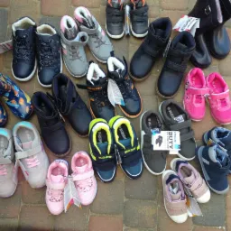 Różnorodny asortyment obuwia dziecięcego i młodzieżowego, w tym botki, tenisówki i kalosze, ułożony na kostce brukowej.