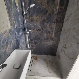 Wykończona łazienka z umywalką nablatową, prysznicem i odpływem liniowym, płytki imitujące marmur w odcieniach szarości i brązu, widoczny fragment niezabezpieczonej ściany przed malowaniem.