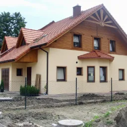 Dwukondygnacyjny dom z beżową elewacją i drewnianą górną częścią, pokryty czerwoną dachówką, otoczony siatką ogrodzeniową i przygotowanym terenem pod ogród.