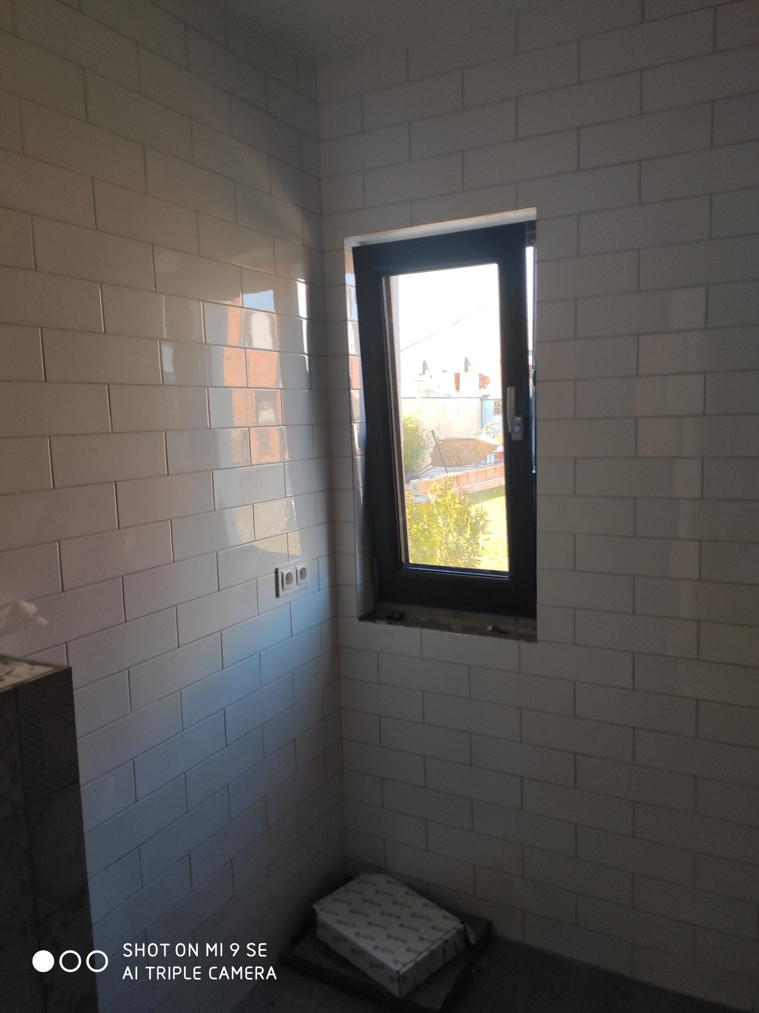 Białe, prostokątne płytki ułożone na ścianie w pomieszczeniu z oknem. Widoczne puszki elektryczne i opakowanie po płytkach na podłodze.