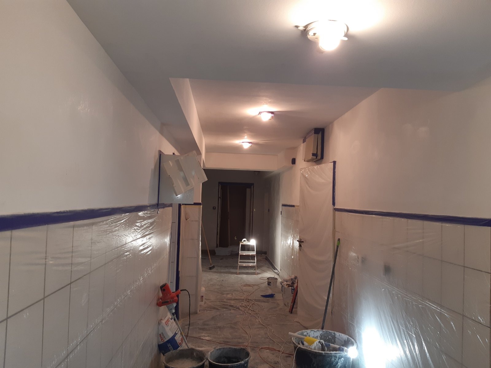 Korytarz w trakcie malowania w Szczecinie, ściany zabezpieczone folią, widoczne wiadra z farbą, mieszadło oraz drabina w głębi.