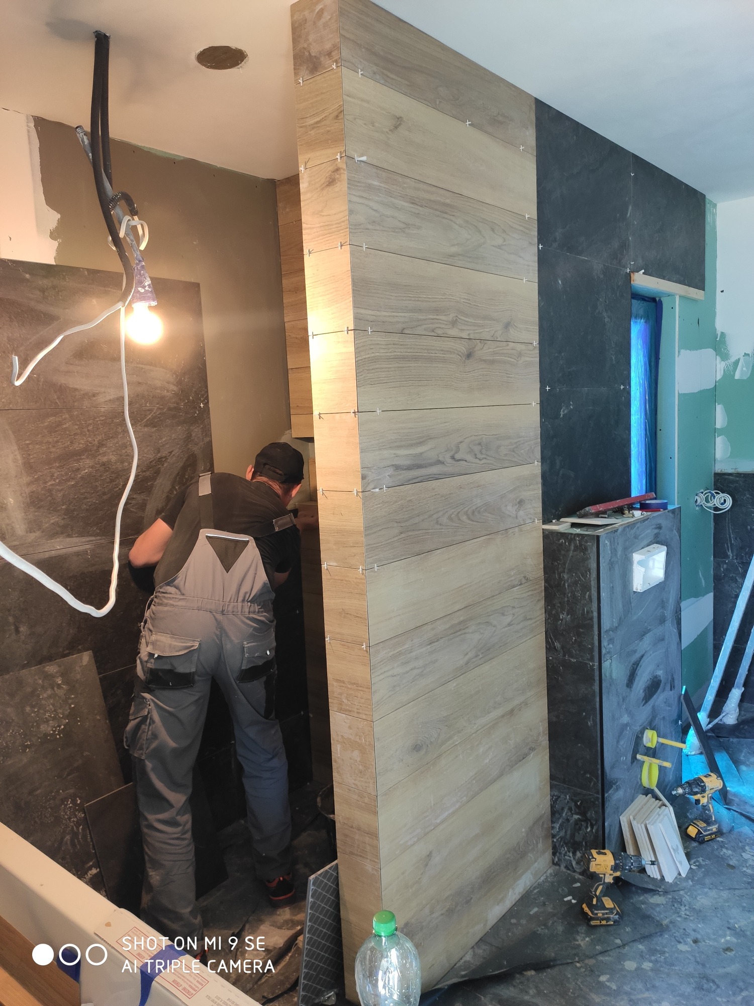 Mężczyzna w szarym kombinezonie montuje drewnopodobne płytki na ścianie w łazience, widoczne krzyżyki dystansowe, obok ściana z ciemnych płytek, narzędzia i przewody elektryczne.