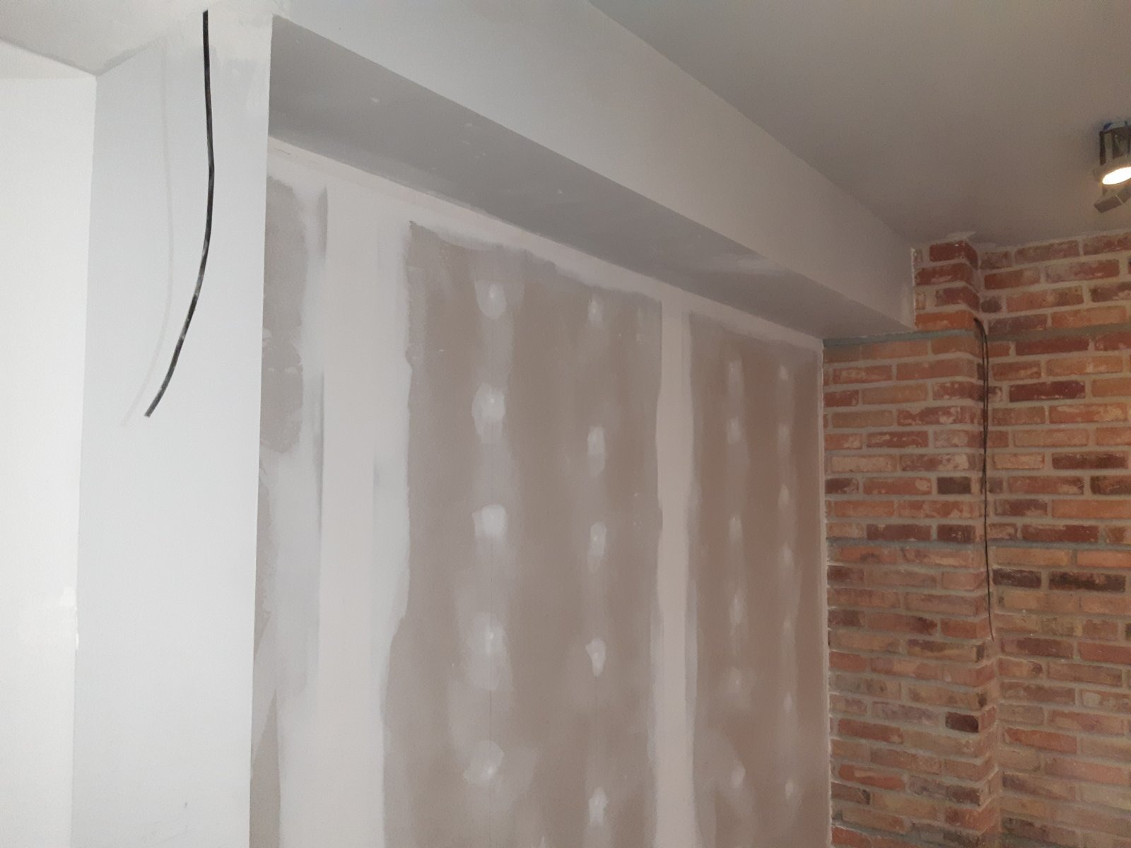 Ściana z płyt kartonowo-gipsowych w trakcie szpachlowania, widoczne łączenia i śruby, fragment ściany z cegły, przewód elektryczny wystający ze ściany.