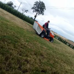 Mężczyzna na pomarańczowej kosiarce samojezdnej tnie trawnik na pochyłym terenie, w tle widoczne drzewa i pochmurne niebo.