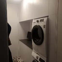 Białe szafki na wymiar z czarnym blatem, pod którymi stoi pralka i suszarka w kolumnie, na podłodze wzorzyste płytki.