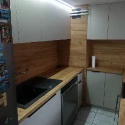 Nowoczesna kuchnia: białe szafki, drewniany blat, czarny zlew. Widoczne magnesy na lod&oacute;wce i subtelne oświetlenie LED pod szafkami g&oacute;rnymi.