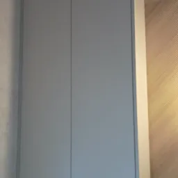 Szara szafka wisząca z dwoma frontami, zamontowana nad podtynkową spłuczką WC w łazience z mozaikową ścianą i teksturowaną tapetą.