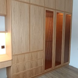 SADEX Meble na wymiar Waldemar Sadowski - Jasna, drewniana szafa wnękowa z ryflowanymi, brązowymi szybami i biurkiem po lewej stronie, na drewnianej podłodze. Widoczny fragment poziomicy.