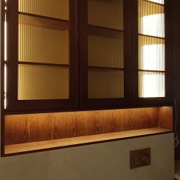 SADEX Meble na wymiar Waldemar Sadowski - Zabudowa meblowa z ryflowanymi szybami i podświetleniem LED nad umywalką. Ciemne drewno, ciepłe światło, nowoczesny design. Widoczne detale armatury.