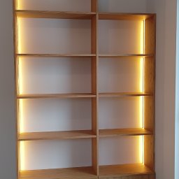 SADEX Meble na wymiar Waldemar Sadowski - Drewniana szafa wnękowa z oświetleniem LED, widok z dołu. Naturalne wykończenie drewna, minimalistyczny design. Na podłodze widoczny odkurzacz.