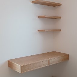 SADEX Meble na wymiar Waldemar Sadowski - Jasne, drewniane półki wiszące i biurko z dwiema szufladami, zamontowane na ścianie w rogu pokoju z panelami podłogowymi. Minimalistyczny design.