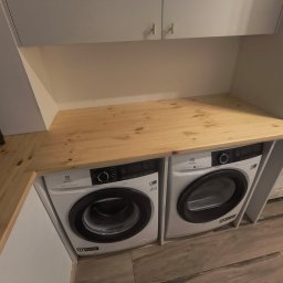 SADEX Meble na wymiar Waldemar Sadowski - Zabudowa pralni z dwoma pralkami Electrolux pod jasnym, naturalnym blatem z drewna sosnowego, z widocznymi sękami. Nad nimi białe szafki z eleganckimi złotymi uchwytami.