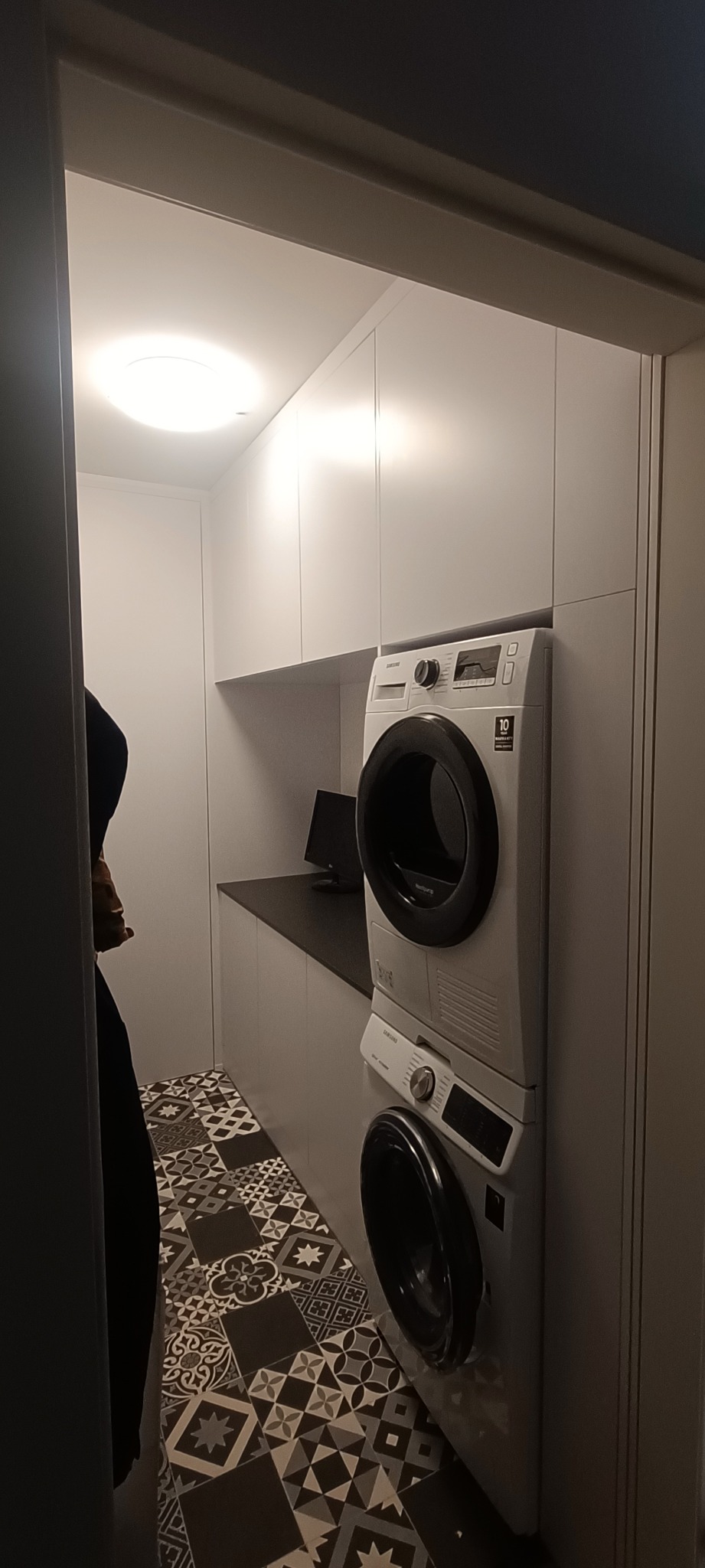 Białe szafy na wymiar z pralką i suszarką w nowoczesnej łazience. Na podłodze wzorzyste płytki. Minimalistyczny design z oświetleniem LED.