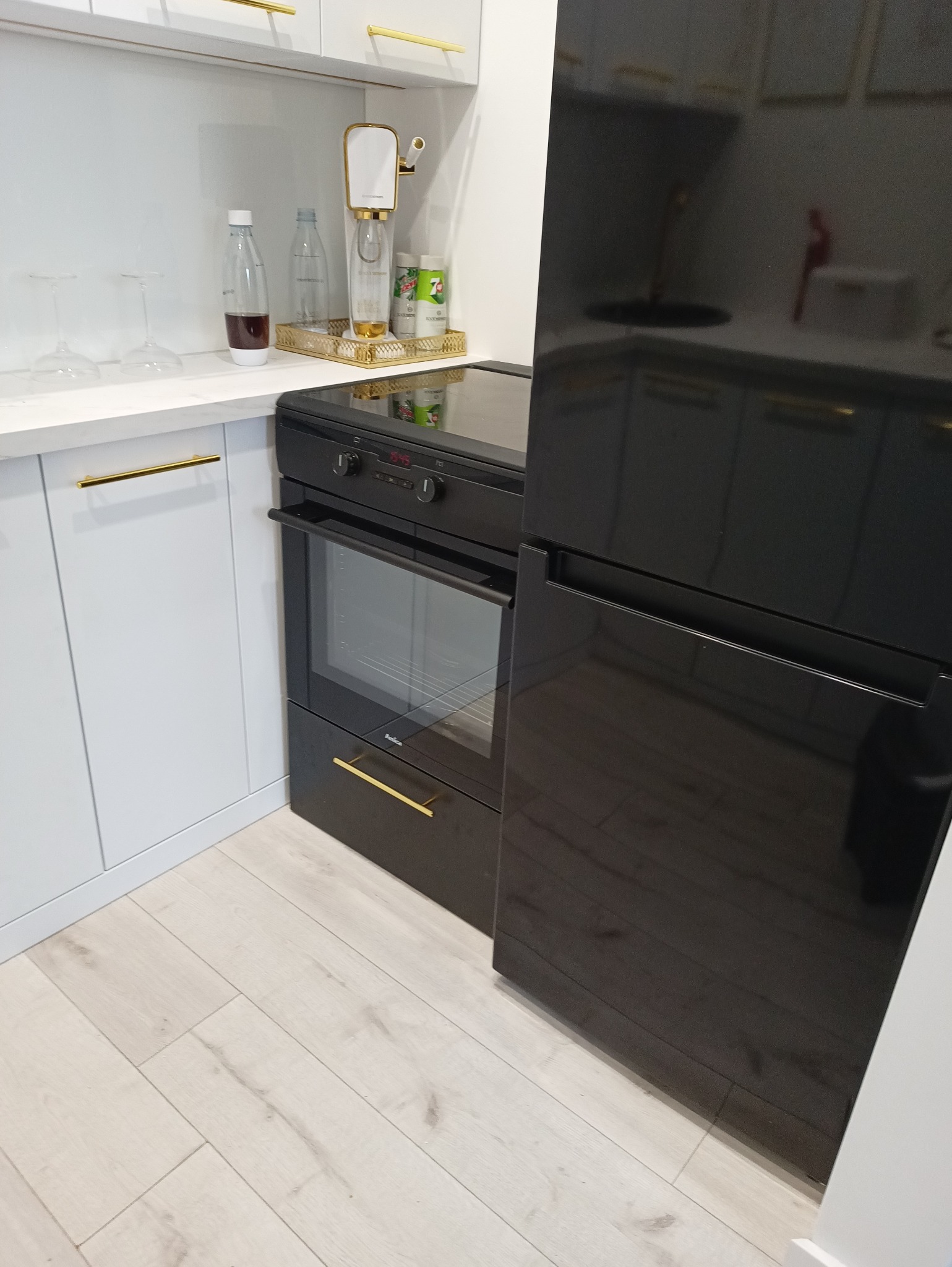 Nowoczesna kuchnia: białe szafki z złotymi uchwytami, czarny piekarnik Amica i lodówka. Na blacie ekspres do wody gazowanej. Jasne panele na podłodze.