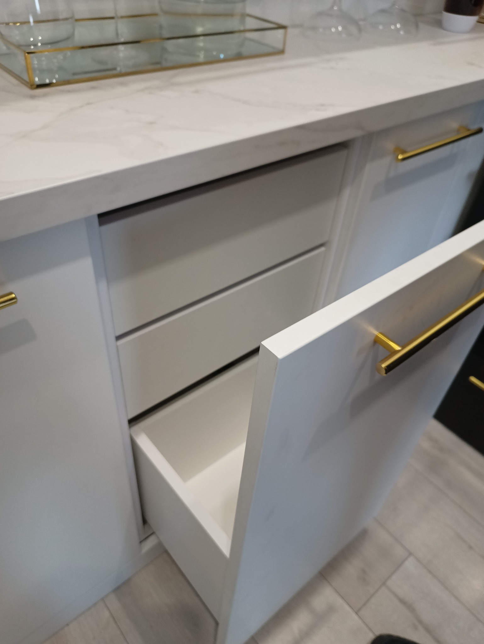 Biała szafka z szufladami i złotymi uchwytami, otwarta szuflada. Blat imitujący marmur. Realizacja z Lubawy.