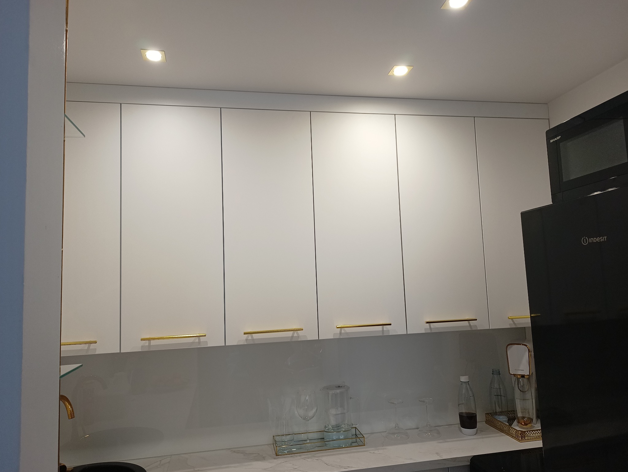 Białe szafki kuchenne pod sufit, z poziomymi złotymi uchwytami, nad blatem z marmurowym wzorem. Widoczny fragment czarnej lodówki i drobne akcesoria kuchenne.
